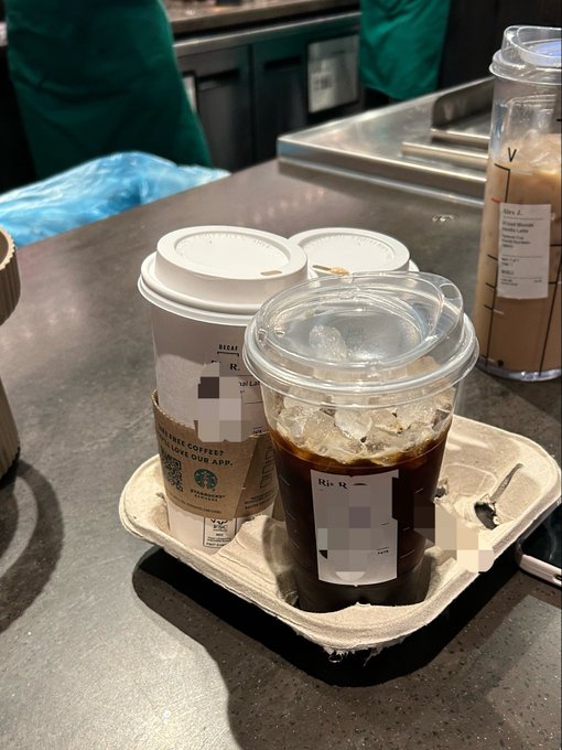 A Starbucks iced coffee in a clear plastic cup sits in a tray alongside another cup, with blurred workers in green aprons and Starbucks equipment visible in the background of the store.