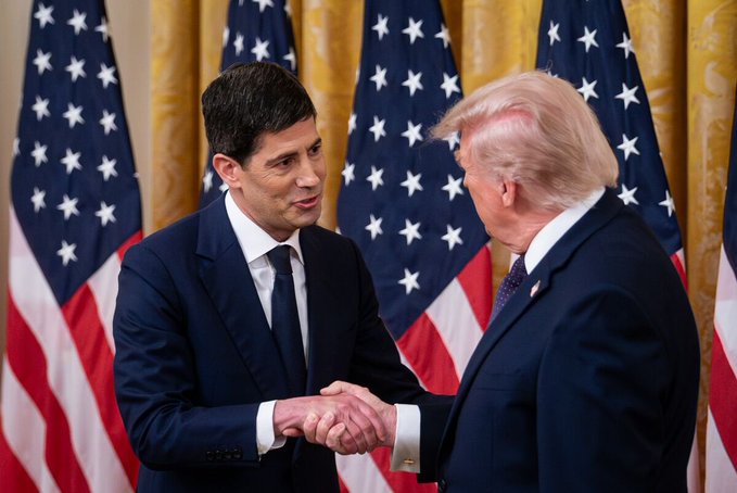 A photograph of two men in dark suits shaking hands in front of several American flags, appearing to be at a formal political or government event.