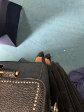 A close-up photo showing a person's feet in black shoes next to a black studded handbag, taken from a slightly elevated angle.