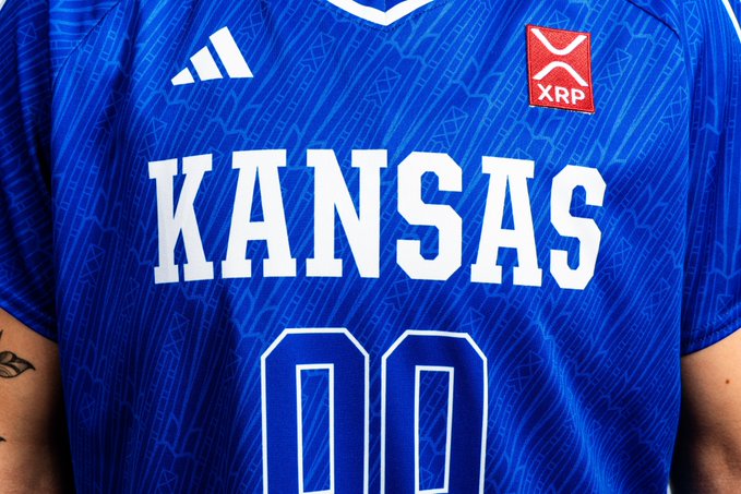 A close-up of a blue Kansas Athletics uniform featuring the Adidas logo, an "XRP" patch from Ripple, and the "KANSAS" team name above the number 00, highlighting the new $XRP branding on the jersey.