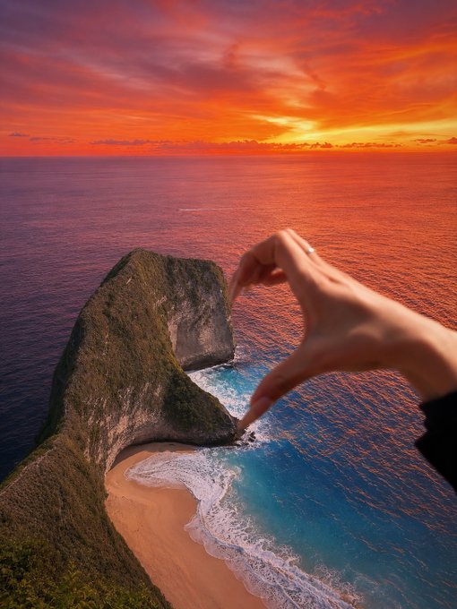 A hand appears to pinch a coastal cliff and beach in a forced perspective shot against a vibrant orange sunset over the ocean.