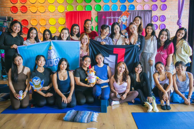 A large group of women pose together in a yoga studio with a colorful polka-dot wall, holding a blue flag with a cartoon character and an 'avalanches' banner.