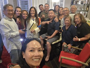 A group selfie of approximately 11 people posing together indoors, with a smiling woman in the foreground and others gathered behind her in what looks like a living room or dining area.