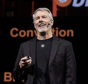 A man with a gray beard smiling while speaking on stage at a convention, with "D" and "Convention" visible in the background.