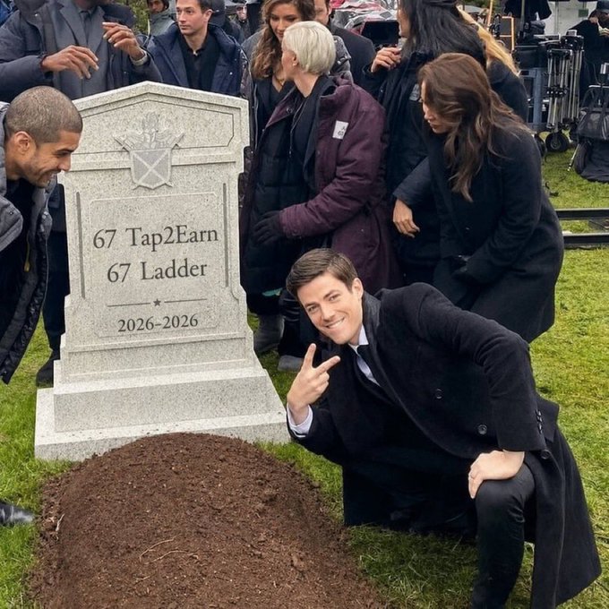 A person in a black suit kneels beside a tombstone reading '67 Tap2Earn, 67 Ladder, 2026-2026,' smiling at the camera as a crowd watches, with a pile of fresh dirt in front.