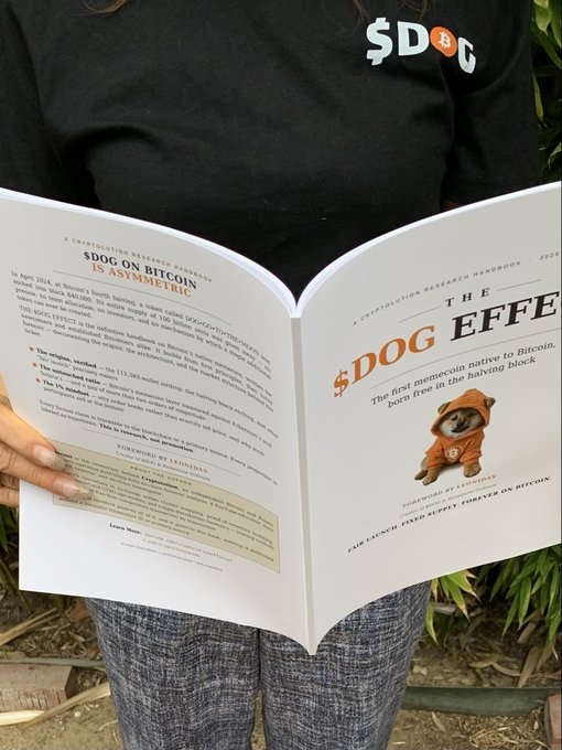 A person wearing a black '$DOG' shirt holds open a research handbook titled '$DOG ON BITCOIN IS ASYMMETRIC' and 'THE $DOG EFFECT,' with $DOG branding and the phrase 'FOR LAUNCH: FIXED SUPPLY FOREVER ON BITCOIN' visible on the right page.