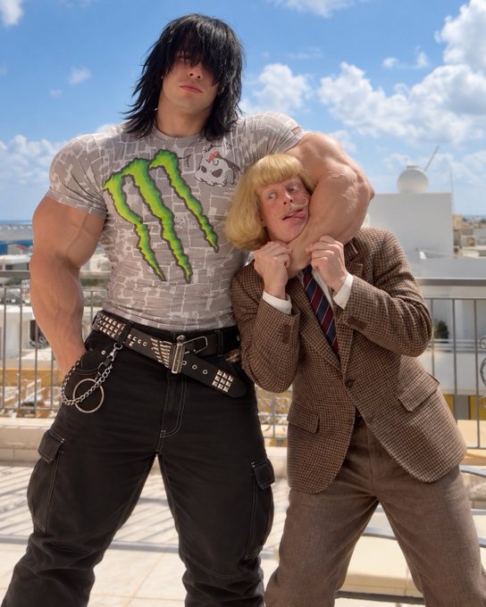 Two people posing together, one wearing a Monster Energy t-shirt with long dark hair, the other in a brown plaid suit with blonde hair, on a rooftop.