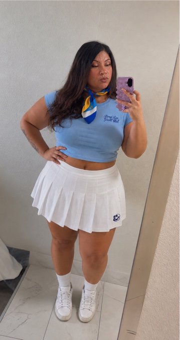 A woman takes a mirror selfie wearing a light blue crop top with a yellow and blue necktie, paired with a white pleated tennis skirt, white socks, and white sneakers, while holding a phone with a purple case. The outfit channels a "schoolgirl" aesthetic that complements her post about embracing her playful "inner lady" vibe as a proud Mutant Ape Yacht Club (MAYC) NFT holder.