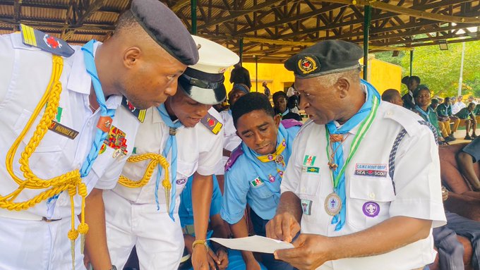 A photograph of uniformed scout/police officers, including one wearing a beret and another in a green cap with a neckerchief, looking at a document together at what appears to be an official event.