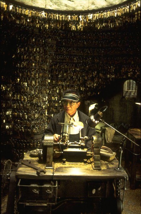 An elderly man with a visor working at a lathe in a workshop filled with hanging keys, from the film 'Hugo'.
