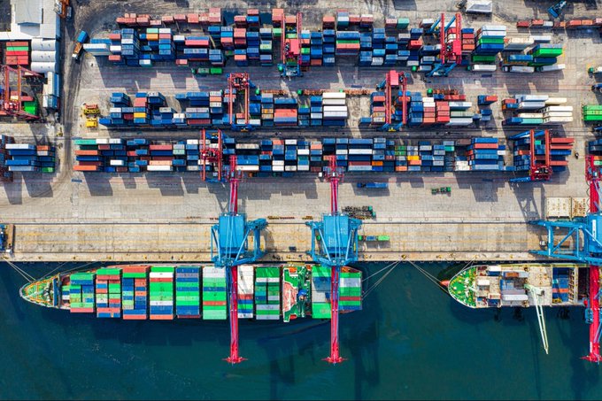 An aerial view of a busy container port shows two cargo ships docked alongside a pier, stacked with thousands of multicolored shipping containers, while massive blue gantry cranes hover above, loading and unloading freight from the dockside stacks. The image of large-scale global trade logistics visually echoes the post's claim that traditional finance moves an enormous $125 trillion in real-world assets annually—Treasuries, funds, credit, and equities—activities that crypto is not yet positioned to handle, despite tokenization efforts.