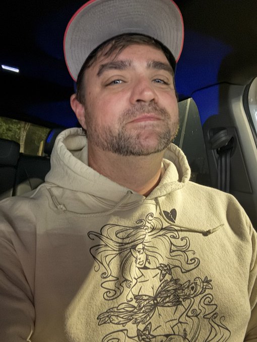 A selfie of a man with a short beard wearing a grey cap and a beige hoodie with a graphic design, sitting in a car.