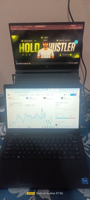 A photo of two laptops stacked, with the top displaying a 'HOLD HUSTLER' trading course by SUCCESS BATTLE, and the bottom showing a crypto trading chart on TradingView, photographed on a realme X7 5G.