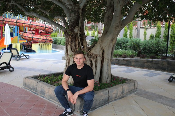 A young man wearing a black t-shirt and dark jeans sitting on a stone planter at the base of a large tree, with a water slide and pool area visible in the background.