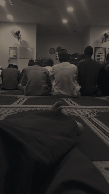 A black-and-white photo shows the backs of several men sitting on a prayer rug in a mosque, with a person's foot visible in the foreground.