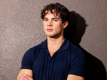 A young man with curly brown hair wearing a navy blue polo shirt, posing with arms crossed against a textured gray wall.
