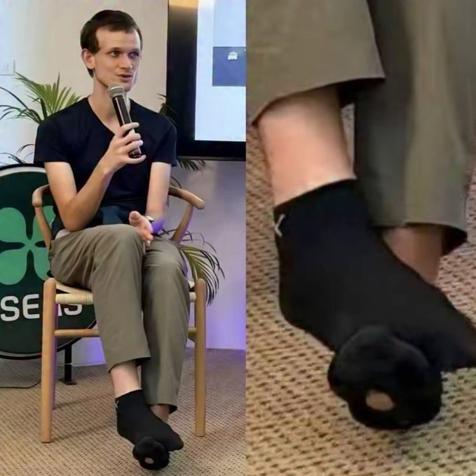 A young man in a black t-shirt and gray pants sits casually in a wooden chair speaking into a microphone at what appears to be a "SEED" event, while wearing visibly worn black toe socks with holes exposing his toes. The close-up shot emphasizes the tattered socks, humorously contrasting with the implied wealth referenced in the caption about crypto success.