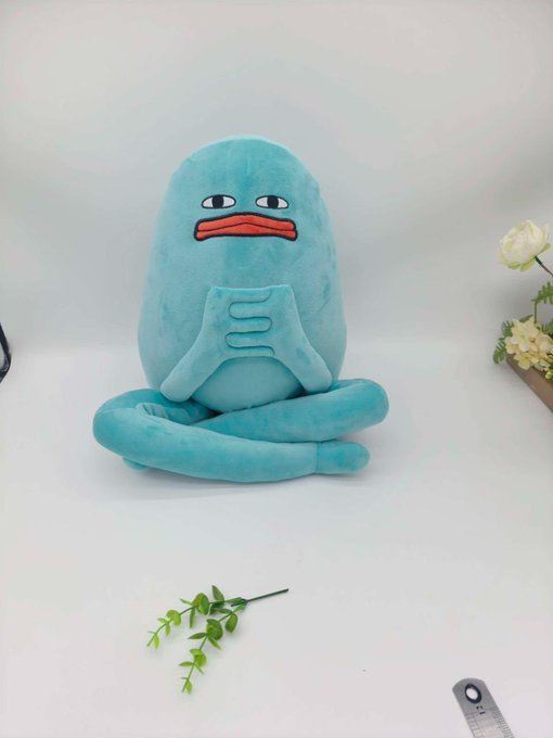 A light blue plush octopus toy with a sad expression sits on a white surface, positioned next to a small sprig of green leaves.