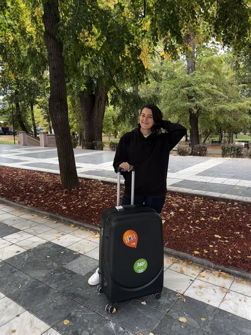 A smiling woman stands outdoors with a rolling suitcase that has round stickers reading 'HiJet' and '360', posing on a paved walkway surrounded by trees.