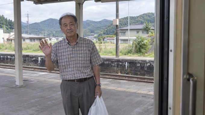 An elderly man in a checkered shirt waves from a rural train platform while holding a plastic bag, set against a backdrop of Japanese countryside houses and mountains. The image's quiet, solitary atmosphere of seeing someone off at a small countryside station pairs with the post's tone of confusion and disbelief, evoking a sense of missing someone or struggling to understand a situation.