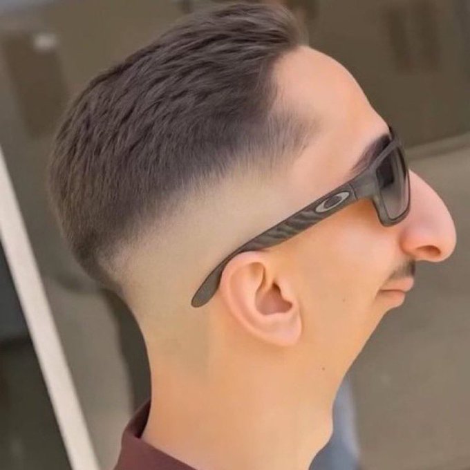 A side profile of a man with a short fade haircut wearing black Oakley sunglasses and a brown shirt.