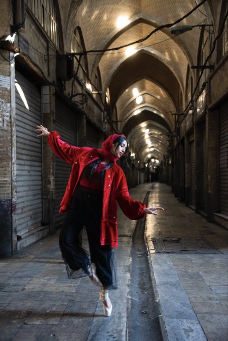 A dancer in a red hooded jacket and black skirt poses on pointe in a dimly lit, arched tunnel or bazaar hallway.