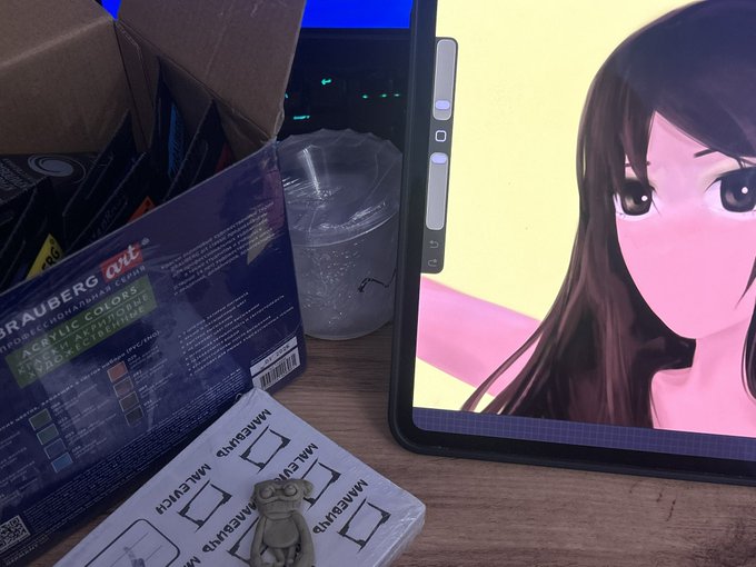 A desk scene showing a tablet displaying anime artwork alongside a 'Trauberg art' acrylic color set box, a small gummy frog figurine, and a 'MAME GUM' package.