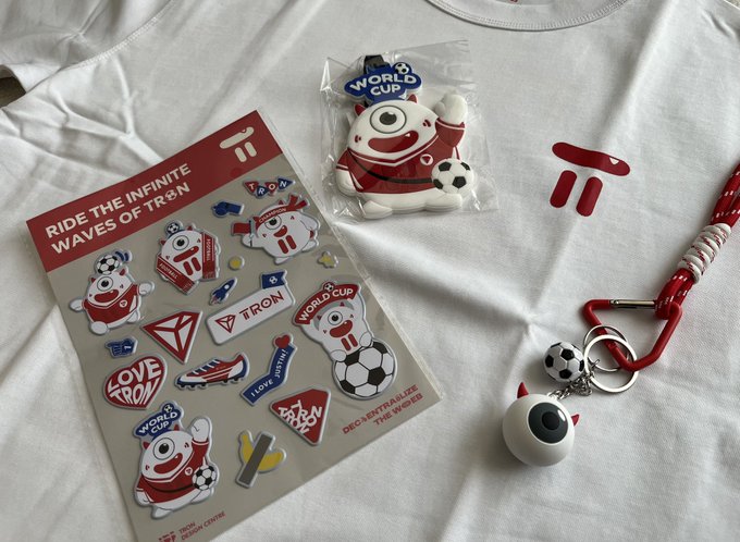 TRON blockchain promotional merchandise laid out on a white t-shirt, including a sticker sheet reading 'RIDE THE INFINITE WAVES OF TRON', a packaged mascot figure, and a keychain with a small character charm.