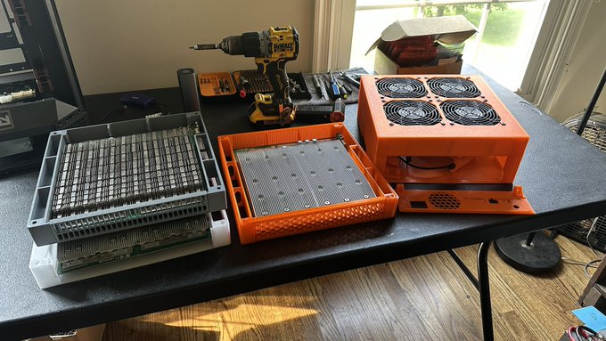 A workbench displaying cryptocurrency mining hardware, including GPU frames stacked in trays and an orange ASIC miner with four cooling fans, alongside a yellow DeWalt drill and tools.