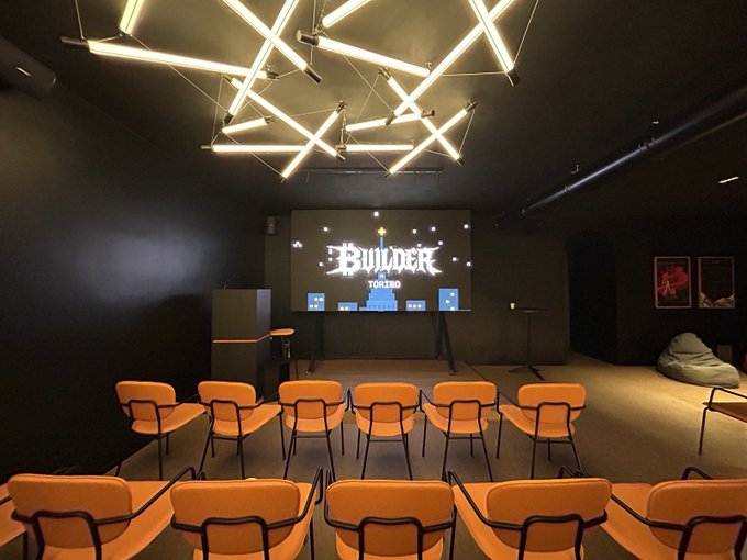 A dimly lit gaming/event room with orange chairs facing a large screen displaying 'BUILDER TOWN', illuminated by a striking geometric neon light fixture on the ceiling.