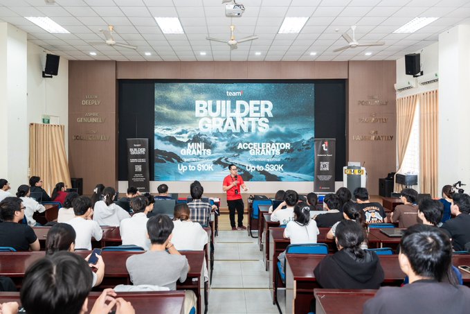 A presentation in a lecture hall where a speaker stands in front of a screen displaying "BUILDER GRANTS" for "MINI GRANTS" up to $10K and "ACCELERATOR GRANTS" up to $30K, while an audience watches from rows of desks.