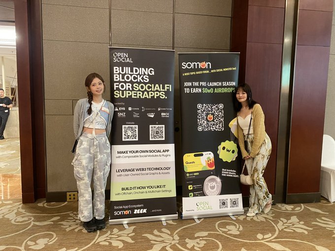 The image shows two women standing next to each other, posing for a photo near some banners or signs. The banners are likely promoting an event or product related to social media or NFTs (Non-Fungible Tokens). One of the women is wearing a yellow shirt and has her arm around the other woman's shoulder. They both appear to be interested in the content displayed on the banners, which could be related to building blocks for social media apps or NFTs.