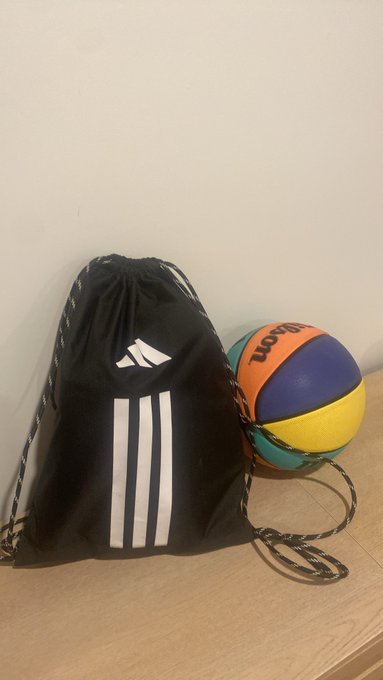 A black Adidas drawstring bag with white stripes sitting next to a multicolored Wilson basketball on a wooden surface.