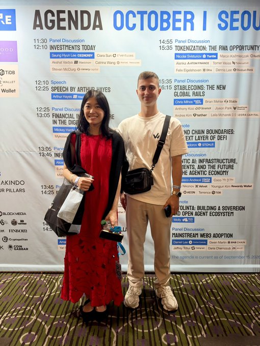 A young woman in a red dress and a young man in a beige t-shirt pose together in front of a conference agenda board listing various panel discussions and speeches, including topics on tokenization, DeFi, and stablecoins.