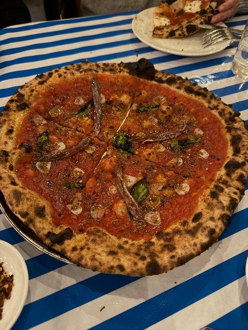 A freshly baked Neapolitan-style pizza topped with tomato sauce, anchovies, and basil sits on a plate atop a blue and white striped tablecloth, with another partially eaten pizza visible in the background.
