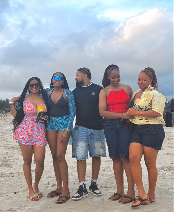 A group of five friends pose together on a sandy beach under a cloudy sky, dressed in colorful beach attire including swimsuits, denim shorts, and sunglasses. The image appears to capture a relaxed, social moment at the Solana Summit Nigeria event.