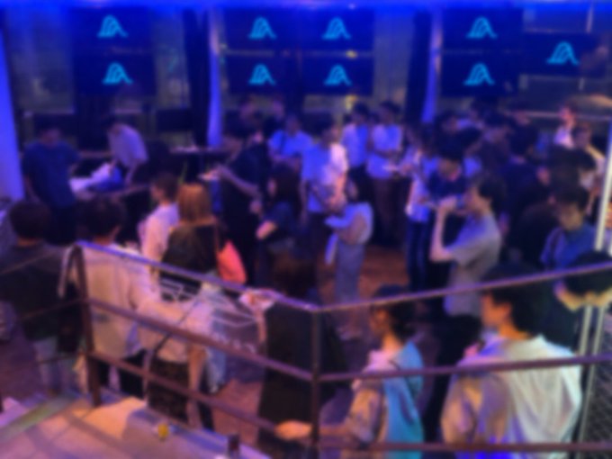 A crowded, dimly lit nightclub or event venue bathed in blue light, featuring multiple screens displaying a triangular logo, possibly a crypto or gaming event.