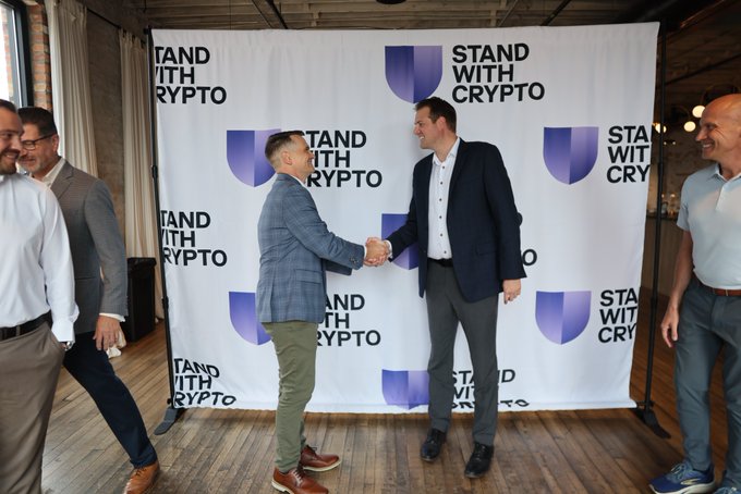 Two men shake hands in front of a "Stand With Crypto" backdrop, joined by other attendees at what appears to be a community roundtable event. The banner's repeated purple logo and the casual indoor setting reflect the grassroots, Main Street focus of the Ohio gathering with Derek Merrin.