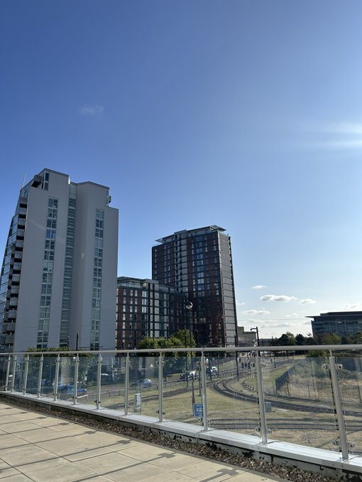A daytime photograph of modern high-rise apartment buildings under a clear blue sky, viewed from an elevated walkway with glass railings, with a small football field visible below.