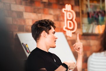 A young man gestures toward a whiteboard with an orange Bitcoin "₿" logo behind him, suggesting an educational or workshop setting. The image complements the post's message about El Salvador's embrace of Bitcoin under President Bukele, illustrating the enthusiasm of a new generation of Salvadorans learning about and engaging with cryptocurrency.