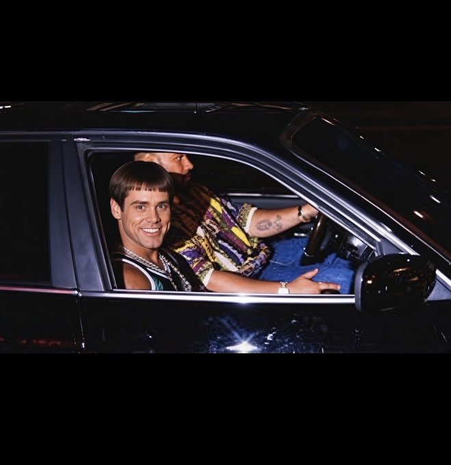 Two young men sitting inside a dark car at night; the passenger is smiling at the camera, while the driver has visible tattoos and is wearing a colorful patterned shirt.