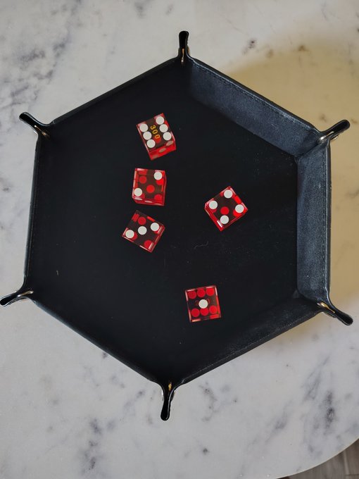 A black hexagonal dice tray resting on a white marble surface, containing five translucent red dice with white pips.