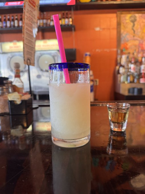A tall highball glass with a blue rim containing a light-colored cocktail and a pink straw sits on a dark bar counter, with a small shot glass of amber liquid nearby.
