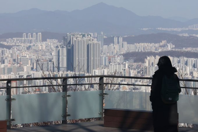 A woman with a backpack stands at a scenic overlook gazing out at a sprawling cityscape of high-rise apartment buildings nestled against misty mountains, visualizing the collective Korean dream of homeownership. The image captures the very subject of the post—tall apartment complexes dominating the urban landscape that represent the ultimate aspiration for many.