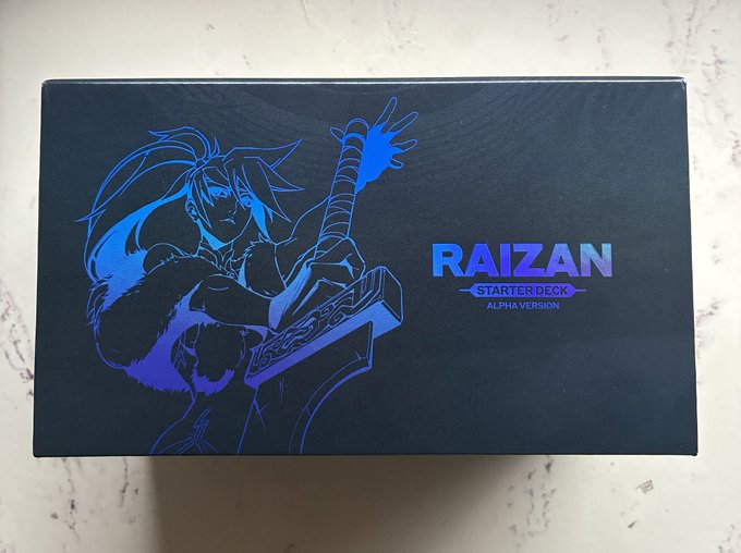A black and blue "RAIZAN STARTER DECK ALPHA VERSION" box featuring artwork of a warrior character wielding a large sword sits on a white surface, which the poster identifies as a sought-after Azuki alpha deck they were lucky to obtain.