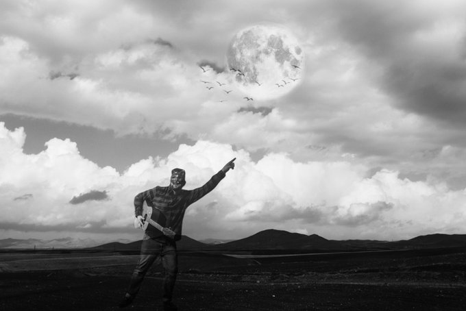A black-and-white photo of a masked figure in a striped sweater pointing toward a large full moon, holding what appears to be an axe, with birds flying across a dramatic cloudy sky.