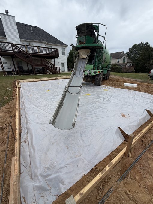 A green cement mixer truck pours concrete down a chute into a wooden-framed foundation lined with a white plastic vapor barrier, with rebar laid nearby in the dirt.