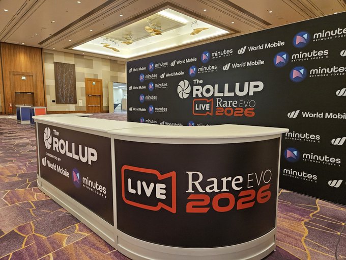 A trade show booth for "The ROLLUP" Live RareEVO 2026 event, powered by World Mobile and minutes Network Token, set up in a conference hall.