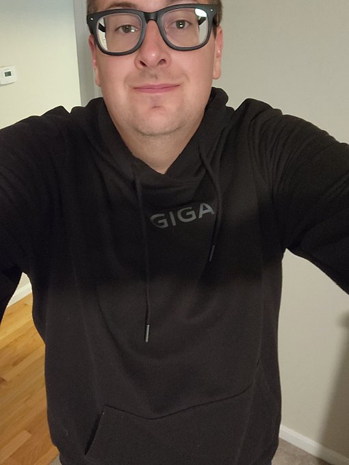 Selfie of a man wearing black-rimmed glasses and a black 'GIGA' hoodie, standing indoors against a plain wall.