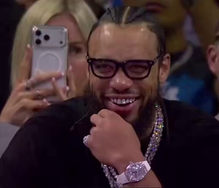 The image features a man wearing glasses, a black shirt, and multiple necklaces with a watch that has diamonds on it. He appears to be smiling at someone taking his picture or recording him. The background shows several people watching something happening behind them, but they are not the main focus of the scene. In the bottom right corner of the image, there is a post about Jupiter Mobile being free to download and how people can use their phones for this purpose.

The man with glasses seems to be enjoying himself as he poses for the camera or someone recording him. The diamonds on his watch add an elegant touch to his overall appearance.